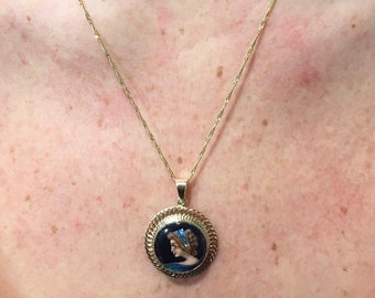 May include: A gold chain necklace with a round pendant featuring a painted portrait of a woman in a blue and white dress. The pendant has a gold filigree border.