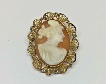 Puede incluir: Un broche de camafeo dorado con un camafeo blanco de perfil de mujer. El camafeo está rodeado por un delicado diseño de filigrana dorada con pequeñas perlas.
