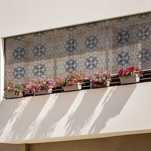 May include: A balcony with a white wall and a patterned window covering. There are five potted plants with pink flowers on the railing.