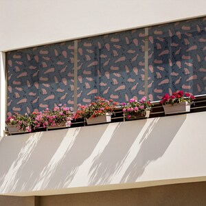 May include: A white building with a balcony featuring a window with pink and blue patterned blinds. The balcony has five potted plants with pink flowers.
