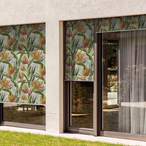 May include: Two windows with white window frames and a white wall behind them. The windows have white sheer curtains and a tropical print roller shade. The roller shade has a pattern of green leaves and orange flowers.