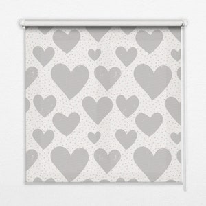 以下が含まれることがあります： 白い背景に大きなグレーのハートが繰り返し描かれたロールスクリーン。小さなピンクの円が散りばめられています。窓やドアに適した、白いローラーメカニズムに取り付けられています。