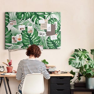 Könnte beinhalten: Eine Frau sitzt an einem Schreibtisch mit einer Korktafel mit tropischen Blattmotiven darüber. Die Korktafel ist mit Fotos und Notizen versehen. Der Text "Thank you" ist auf einer der Notizen sichtbar. Der Schreibtisch ist aus Holz und hat einen schwarzen Metallrahmen. Auf dem Schreibtisch steht eine Topfpflanze und an der Wand hängt eine Uhr.