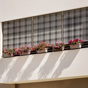 May include: A white building with a balcony featuring a window with a plaid patterned shade and several potted plants with pink and red flowers.