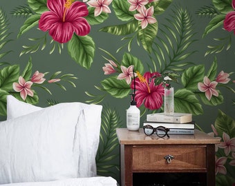 Puede incluir: Un dormitorio con un papel tapiz floral tropical verde con flores rosas y rojas. Una cama blanca con un edredón blanco y una almohada blanca está en primer plano. Una mesita de noche de madera con un cajón y una cámara en la parte superior está al lado de la cama. La mesita de noche también tiene una pila de libros y un par de gafas en la parte superior.