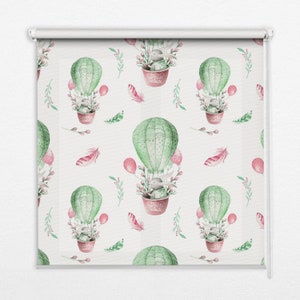 Op de afbeelding: Een witte rolgordijn met een herhalend patroon van groene luchtballonnen met roze accenten. Elke ballon heeft een klein grijs konijn erin en is omringd door roze en groen gebladerte.