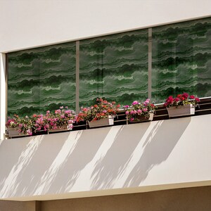 May include: A white building with a balcony featuring a row of potted pink and red flowers. The balcony has a glass railing and a green patterned window behind the flowers.