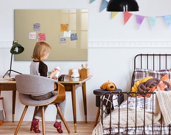 May include: A child sits at a desk with a beige magnetic dry erase board on the wall behind them. The board has colorful sticky notes with handwritten text on it. The child is holding a small white container. The desk has a wooden top and black metal legs. There is a green lamp on the desk. There is a bed with a black metal frame and a plaid comforter in the background.