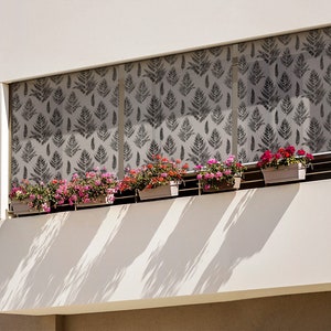 May include: A white building with a balcony featuring a set of gray patterned window shades and several potted plants with pink flowers. The balcony is shaded by the window shades.