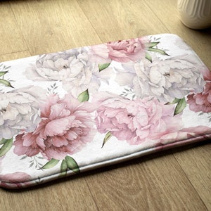 Può includere: Un tappetino da bagno bianco con un motivo floreale di peonie rosa e bianche. Il tappetino ha una texture morbida e soffice ed è perfetto per aggiungere un tocco di eleganza a qualsiasi bagno.