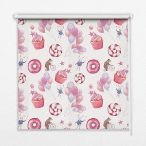 以下が含まれることがあります： ピンクと白のパターンが施された白いローラーブラインド。パターンには、カップケーキ、ドーナツ、星、風船、ウサギが描かれています。