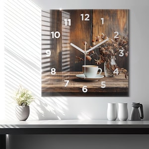 Op de afbeelding: Een witte klok met zwarte Romeinse cijfers op een houten achtergrond. De wijzers van de klok zijn wit en wijzen naar 3:00 uur. Een kopje koffie staat op een schoteltje voor een vaas met gedroogde bloemen.