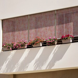 May include: A white building with a balcony featuring a row of potted pink and red flowers. The balcony has a white railing and a patterned window covering.
