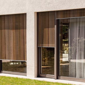 May include: Two large windows with wooden slat blinds. The windows are on a white wall with a light gray stucco finish. The windows are framed in dark brown metal.