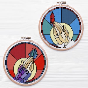 Könnte beinhalten: Zwei runde Kreuzstich-Stickrahmen. Jeder hat ein Design im Glasmalerei-Stil mit Händen, die eine Feder halten. Einer hat eine rote Feder und einen blauen Hintergrund, der andere eine blaue Feder und einen roten Hintergrund. Die Reifen sind vor einem weißen Holzhintergrund angeordnet.