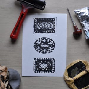 May include: A white sheet with three black linocut prints of insects and animals. The prints feature a moth, a cheetah-like creature, and a beetle. A red and black print roller and other printmaking tools are visible.