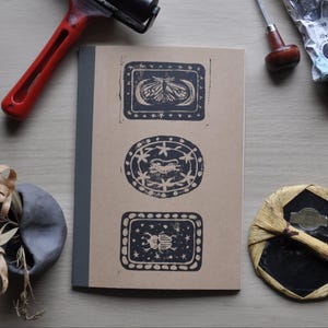 Peut inclure: Un carnet marron avec trois motifs noirs imprimés au bloc : un papillon de nuit, une créature et un scarabée. L'image comprend des outils de gravure, tels qu'une roulette rouge et des outils de sculpture, suggérant un thème de gravure.