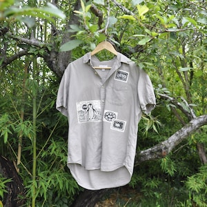 Op de afbeelding: Een lichtgrijs overhemd met korte mouwen en verschillende patches. Eén patch toont een pinguïn met het woord "STOP". Het overhemd hangt aan een houten hanger tegen een groene, bladerrijke achtergrond.