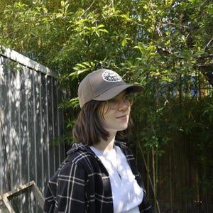 May include: A brown baseball cap with a circular patch design. The cap is worn with a white t-shirt and a black and white plaid shirt. The background features green foliage and a wooden fence.