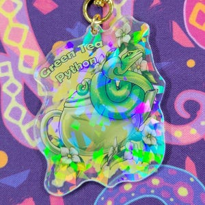 Könnte beinhalten: Ein klarer Acryl-Schlüsselanhänger mit einem grünen Schlangen-Design, das sich um eine Teekanne windet, mit dem Text "Green Tea Python". Der Schlüsselanhänger hat einen goldenen sternförmigen Verschluss und holografische Details.
