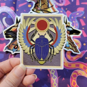 May include: A sticker featuring a blue scarab beetle with gold accents, surrounded by golden wings and a red sun. The sticker has a vintage aesthetic and is on a white background.