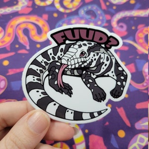 May include: A white and black lizard sticker with a pink tongue sticking out. The sticker has the word "FUUD?" written in pink on it.