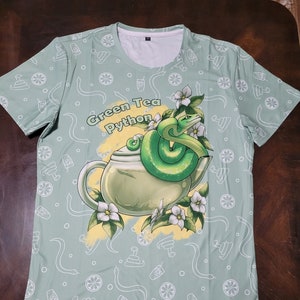 Peut inclure: Un t-shirt vert avec un fond vert clair et un motif blanc de citrons, de serpents et de boissons. Le t-shirt présente un graphique d'un serpent vert enroulé autour d'une théière avec le texte "Green Tea Python".