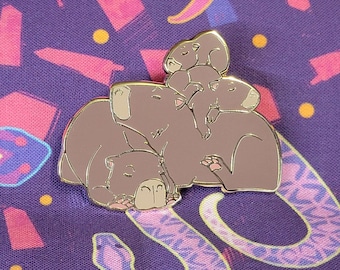May include: A gold-tone enamel pin featuring a group of four capybaras huddled together. The capybaras are brown and have white markings on their faces. The pin is on a purple background with a colorful snake pattern.