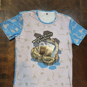 Peut inclure: Un t-shirt blanc avec un motif bleu et blanc. Le t-shirt présente un graphique d'un serpent buvant dans un verre avec le texte "Butterball Python".