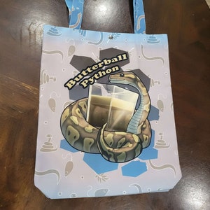 以下が含まれることがあります： 白い背景に蛇、ネズミ、その他の小さなオブジェクトのパターンが描かれた薄い青色のトートバッグ。バッグには、液体が入ったグラスに巻き付いた蛇の大きなグラフィックがあり、黄色の文字で「Butterball Python」と書かれています。