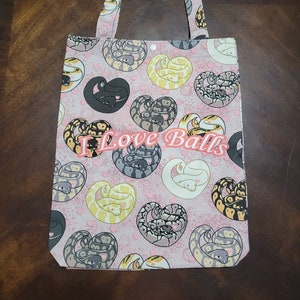 May include: A pink canvas tote bag with a white and pink heart pattern featuring cartoon snakes. The text "I Love Balls" is printed in orange on the bag.