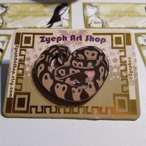 Puede incluir: Una serpiente marrón y negra con forma de corazón con un corazón rosa en la cabeza. La serpiente está sobre un fondo dorado con el texto "Zyepth Art Shop" y un nombre de perfil en las redes sociales.