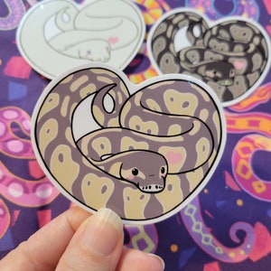 May include: A set of three heart-shaped stickers featuring cartoon snakes. The stickers are printed on white glossy paper and have a shiny finish. The snakes are curled up in a heart shape and have cute faces. The snakes are different colors: one is white, one is brown and black, and one is light brown and beige.