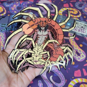 May include: A red and yellow enamel pin featuring a humanoid figure with a lobster tail and claws. The figure is wearing a crown and has a fierce expression.