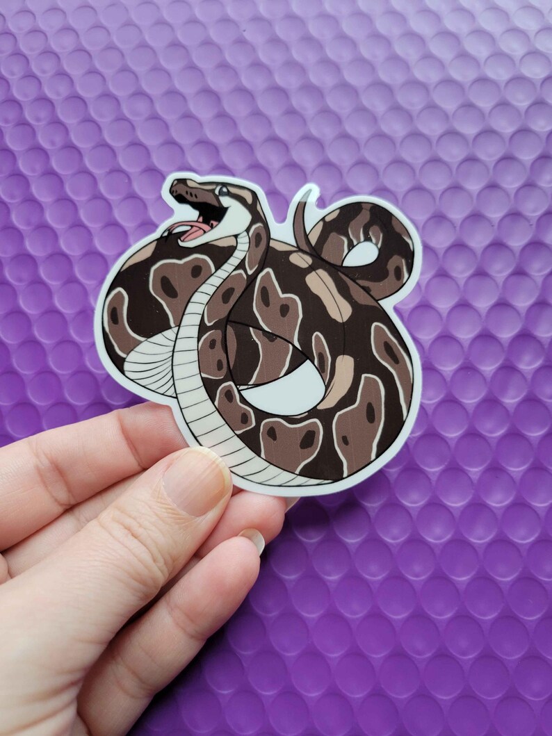 Puede incluir: Una pegatina de una serpiente marr&oacute;n y blanca con un dise&ntilde;o estampado. La serpiente est&aacute; enroscada con la boca abierta, como si fuera a atacar.