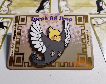 May include: A gray and white enamel pin of a yellow-crested cockatiel with its wings spread. The pin is on a brown card with gold accents and the text "Zueph Art Shop" and "ZuephArtShop.etsy.com".