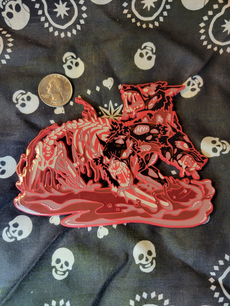 May include: A red enamel pin depicting a three-headed wolf with a skeletal body, set against a red background. The wolf's heads are snarling and have sharp teeth. The pin is a detailed and intricate design.