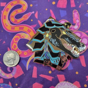 May include: A detailed metal pin in the shape of a stylized dragon head, featuring black, gold, and teal accents. The pin is placed on a purple fabric with a snake pattern, next to a US quarter.