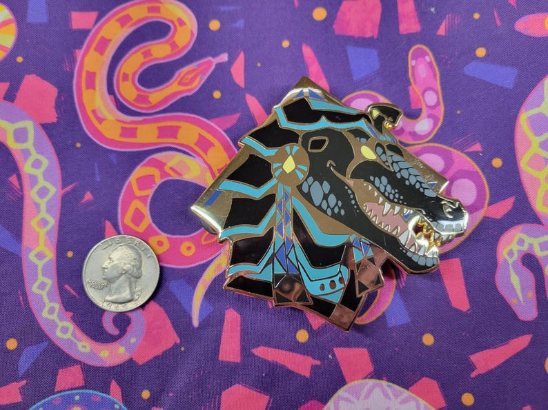 May include: A detailed metal pin in the shape of a stylized dragon head, featuring black, gold, and teal accents. The pin is placed on a purple fabric with a snake pattern, next to a US quarter.