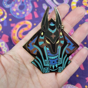 May include: A gold-colored Anubis pin with black, blue, and brown accents. The pin features a stylized depiction of the Egyptian god Anubis, with a geometric design. The pin is held in a hand against a purple background with snake patterns.