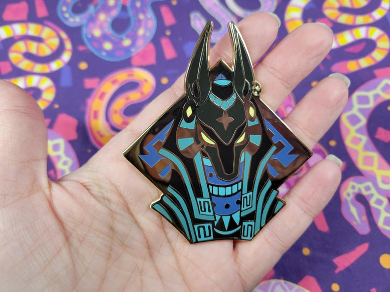May include: A gold-colored Anubis pin with black, blue, and brown accents. The pin features a stylized depiction of the Egyptian god Anubis, with a geometric design. The pin is held in a hand against a purple background with snake patterns.