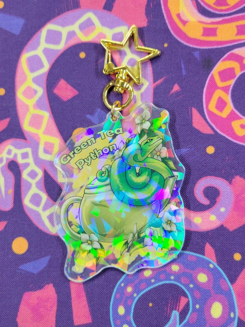 K&ouml;nnte beinhalten: Ein klarer Acryl-Schl&uuml;sselanh&auml;nger mit einem gr&uuml;nen Schlangen-Design, das sich um eine Teekanne windet, mit dem Text "Green Tea Python". Der Schl&uuml;sselanh&auml;nger hat einen goldenen sternf&ouml;rmigen Verschluss und holografische Details.