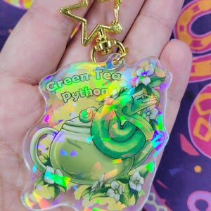 K&ouml;nnte beinhalten: Ein klarer Acryl-Schl&uuml;sselanh&auml;nger mit einem gr&uuml;nen Teekannen- und Schlangen-Design, mit dem Text "Green Tea Python". Er hat einen goldfarbenen sternf&ouml;rmigen Verschluss und eine Kette. Das Design beinhaltet wei&szlig;e Blumen und einen holografischen Effekt.