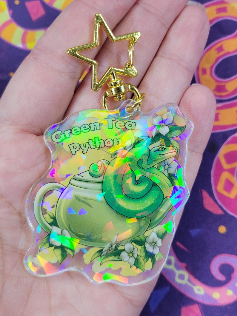 K&ouml;nnte beinhalten: Ein klarer Acryl-Schl&uuml;sselanh&auml;nger mit einem gr&uuml;nen Teekannen- und Schlangen-Design, mit dem Text "Green Tea Python". Er hat einen goldfarbenen sternf&ouml;rmigen Verschluss und eine Kette. Das Design beinhaltet wei&szlig;e Blumen und einen holografischen Effekt.