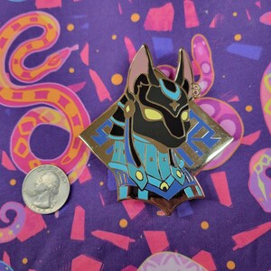 May include: A gold-colored Anubis pin with black, blue, and yellow accents. The pin is in the shape of a diamond with the Anubis head in the center. The pin is on a purple background with a quarter for size comparison.