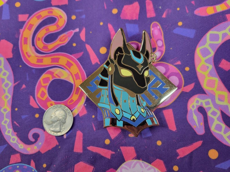 May include: A gold-colored Anubis pin with black, blue, and yellow accents. The pin is in the shape of a diamond with the Anubis head in the center. The pin is on a purple background with a quarter for size comparison.