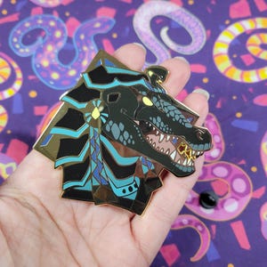 May include: A detailed enamel pin featuring a stylized dragon head with gold, black, and teal accents. The dragon has an open mouth with visible teeth and a gold tooth. The pin is held against a purple background.