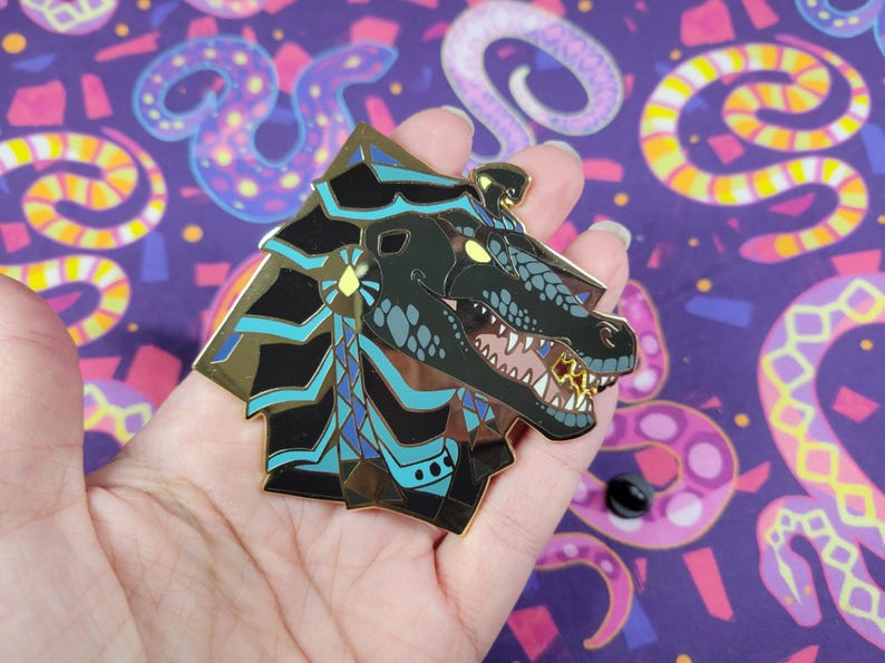 May include: A detailed enamel pin featuring a stylized dragon head with gold, black, and teal accents. The dragon has an open mouth with visible teeth and a gold tooth. The pin is held against a purple background.