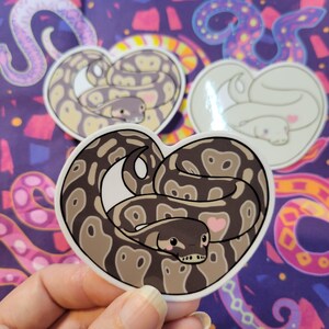May include: A sticker of a brown and black snake curled up in a heart shape. The snake has a pink heart on its side and a cute cartoon face. The sticker is on a purple background with a pattern of colorful snakes.
