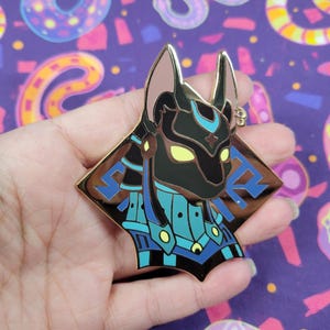 May include: A gold-colored Anubis pin with black, blue, and yellow accents. The pin features a detailed depiction of the Egyptian god Anubis, with a stylized design and a diamond-shaped border. The pin is likely made of metal and has a shiny finish.